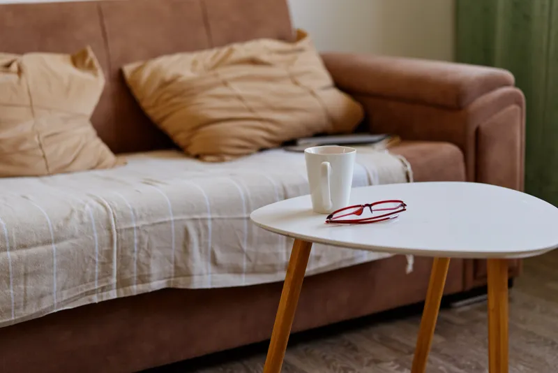 Cozy livingroom scene shows a round white coffee table with a cup and red glasses beside a beige sofa and plush cushions, conveying a calm home mood.