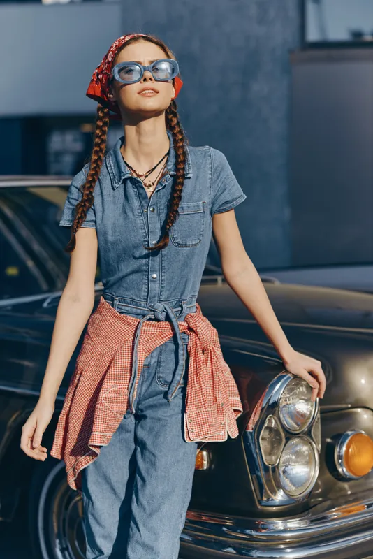 stylish woman in denim outfit with braided hair and sunglasses posing near vintage car under bright sunlight on urban street