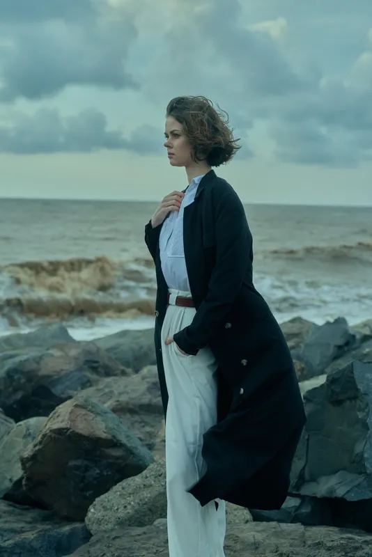 Contemplative woman on a rocky coast facing the sea, wearing a long coat and white ensemble, wind lifts hair as stormy clouds gather
