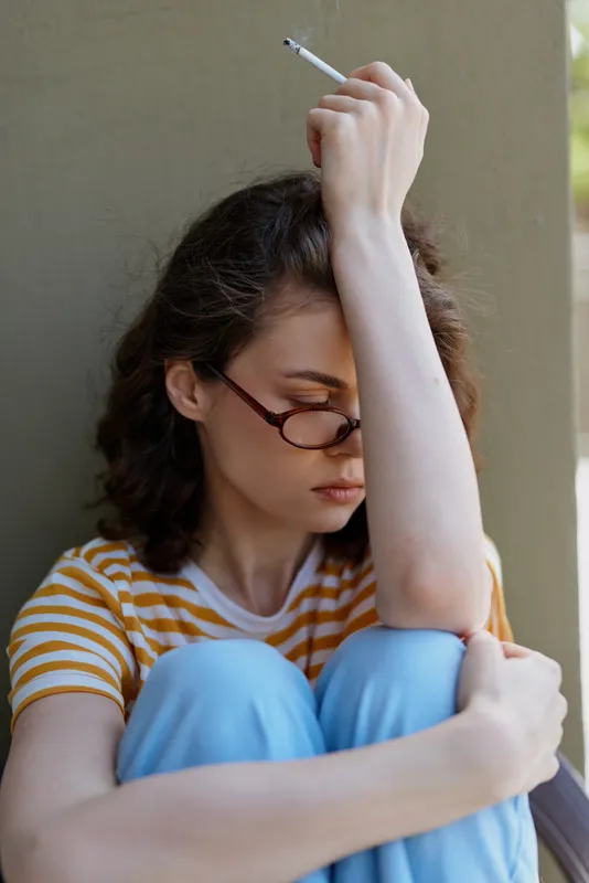 A pensive woman sits against a wall with one arm raised, cigarette in hand, wearing a striped shirt and light pants, conveying a moody urban moment and quiet contemplation.