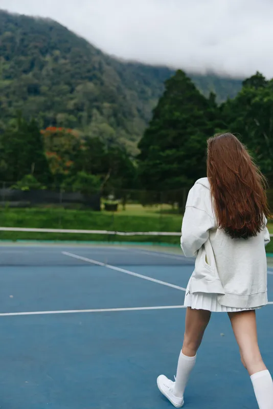 Young woman in a white outfit walking on a tennis court, surrounded by lush green mountains under a cloudy sky, showcasing fitness and an active lifestyle