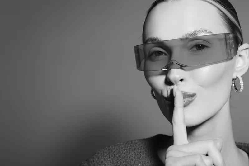 Close-up of a fair skinned white woman with light hair wearing transparent glasses and making a silence gesture on a grey background.