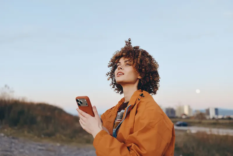 Woman lifestyle outdoors, smartphone in hand, portrait of curly hair, orange jacket, standing near seaside shore, looking upward in wind, warm golden light, relaxed pose, soft sky
