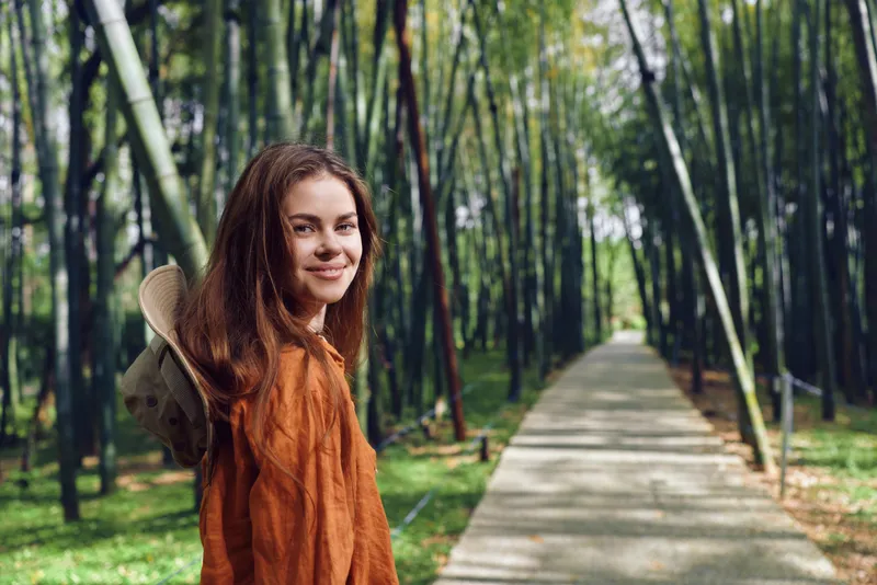 Woman with backpack and smile on a concrete path through a bamboo forest, travel mood and hiking adventure. Casual portrait, green nature trail and peaceful outdoor scene.