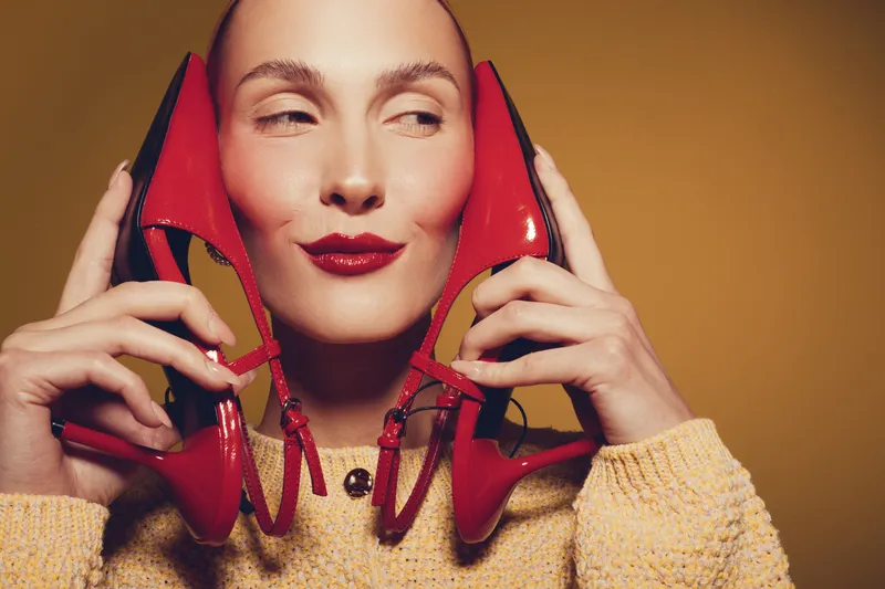 Woman with red lipstick holds red high heel shoes near face, wearing yellow sweater. Studio portrait with warm lighting on brown background. Concept of fashion, style, and beauty.