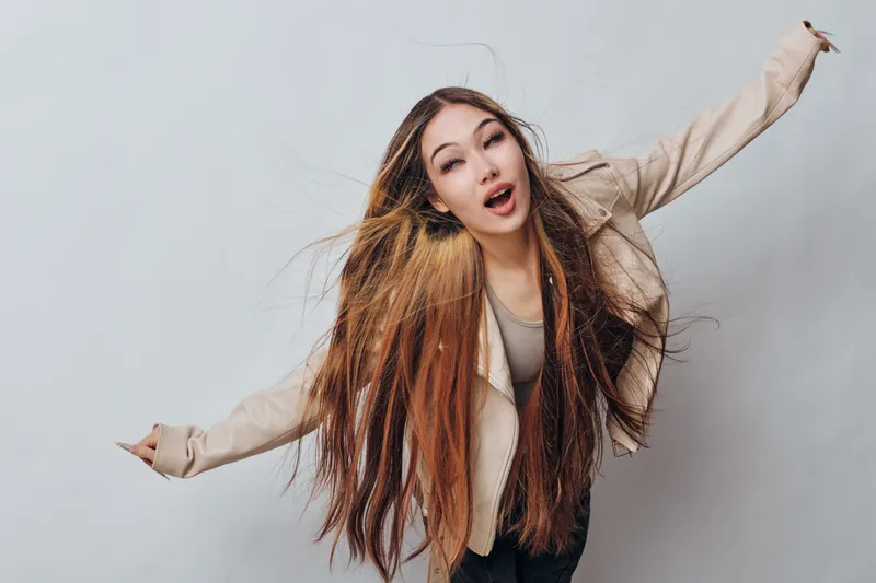 Young woman with long hair and light jacket posing with arms outstretched on gray background, expressing joy and freedom with natural makeup and casual style.