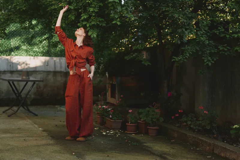 A young woman in a rust jumpsuit raises her arm in a sunlit garden, conveying freedom, curiosity and a playful mood