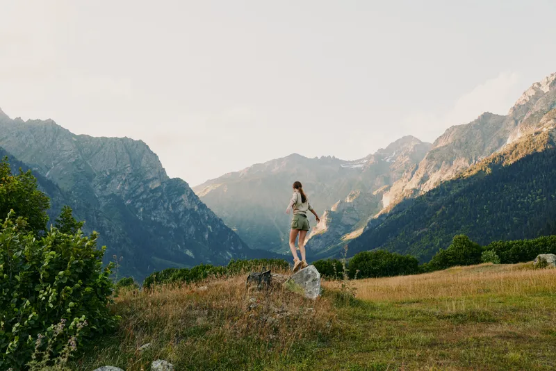 A woman hiking along a grassy trail in a mountain landscape, embracing outdoor solitude and the refreshing air of nature. The scene suggests calm and wellness on the move.