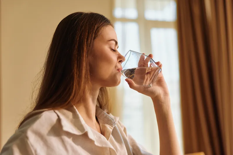 Woman drinking water from glass at home in soft natural light, portrait capturing a relaxed morning routine, hydration and wellness with casual shirt and calm expression.