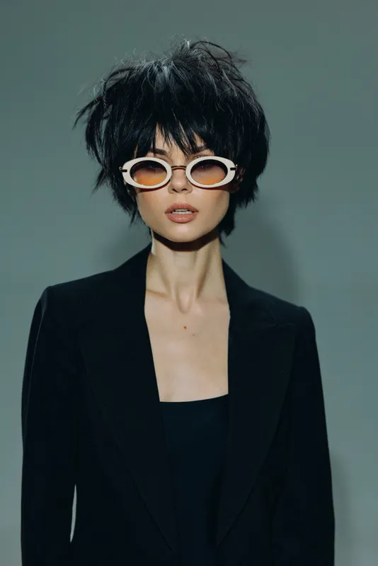 Stylish woman with messy black hair wearing white sunglasses and a black blazer posing confidently indoors.