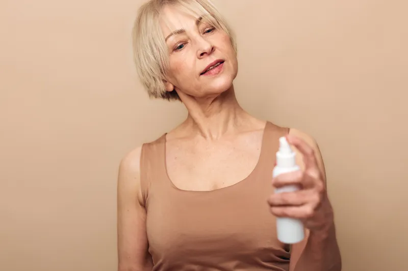 Mature woman with short blonde hair sprays facial mist on her face, wearing beige tank top. Concept of skincare, hydration, beauty routine, and self-care for aging skin.