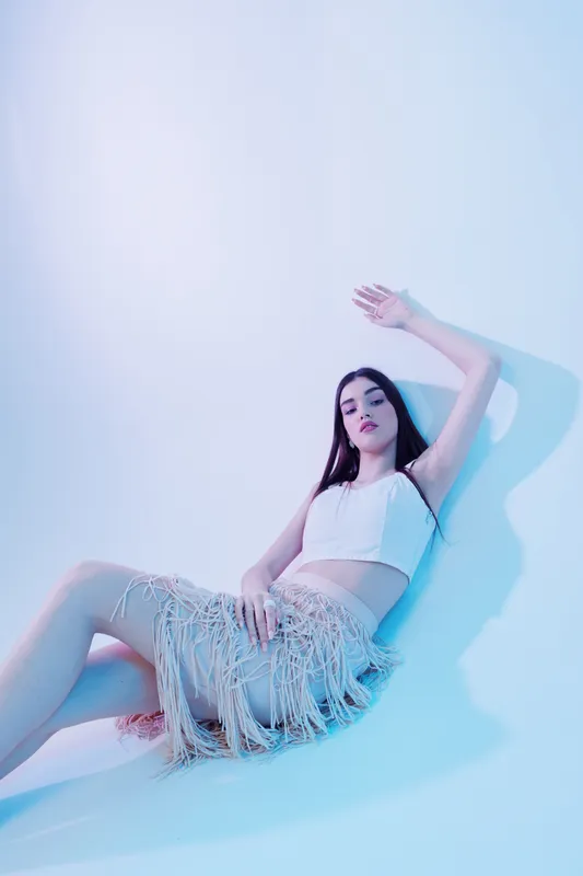 Young woman in a stylish white crop top and fringed skirt poses against a light blue background. Concept of woman beauty fashion with soft lighting and modern style.