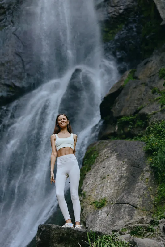 young woman in white sportswear standing confidently by waterfall outdoors, natural scenery, fitness, serene atmosphere, active lifestyle