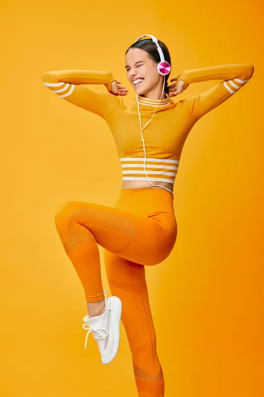 Female fitness enthusiast wearing orange activewear performs a lively dance pose, exuding energy and athletic confidence in a vibrant studio backdrop. Her headphones underline a dynamic, upbeat