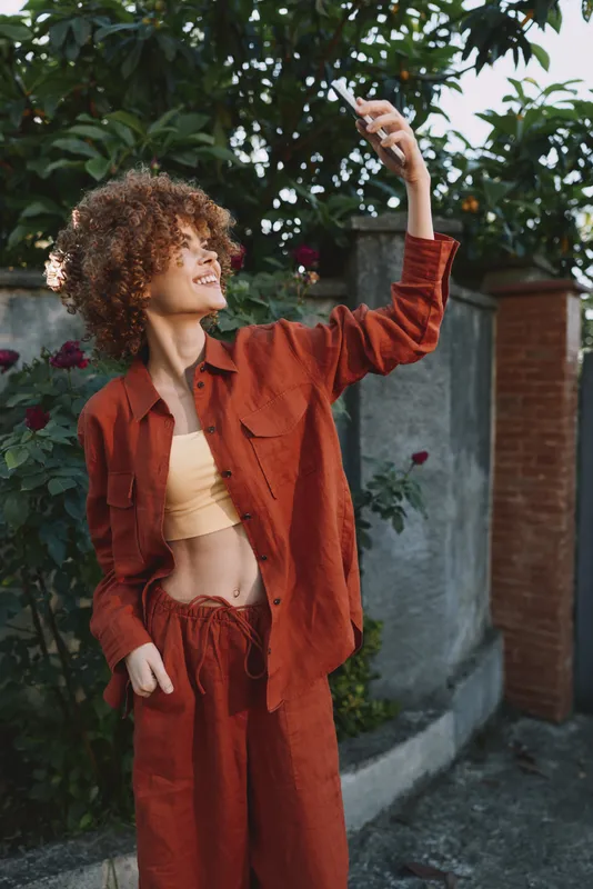 Curly haired woman in a burnt orange tracksuit strikes a confident pose outdoors, capturing a selfie against a brick and floral backdrop. Fashion, lifestyle, and self expression