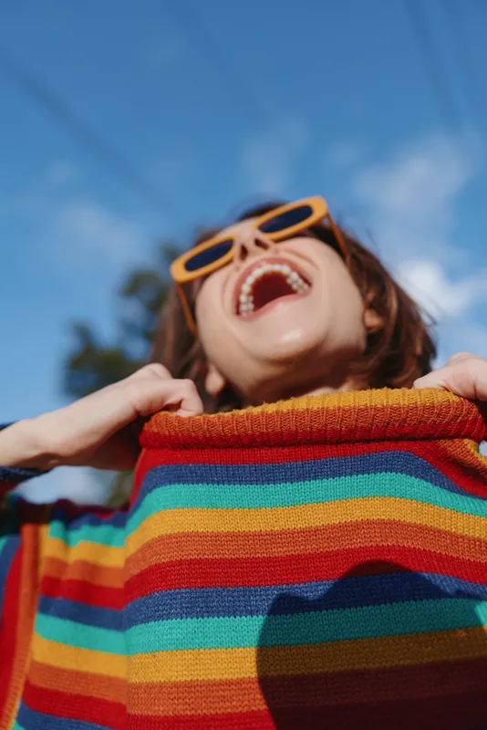 Young woman in a rainbow sweater laughing with yellow sunglasses under a blue sky in a tropical outdoor setting, joyful lifestyle portrait for travel and casual fashion.