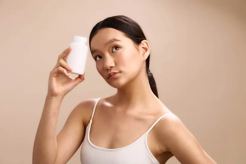 Asian woman holds a white bottle in a minimalist studio, radiating a fresh, confident mood. The clean setting highlights wellness and self care, inviting skincare routine ideas.