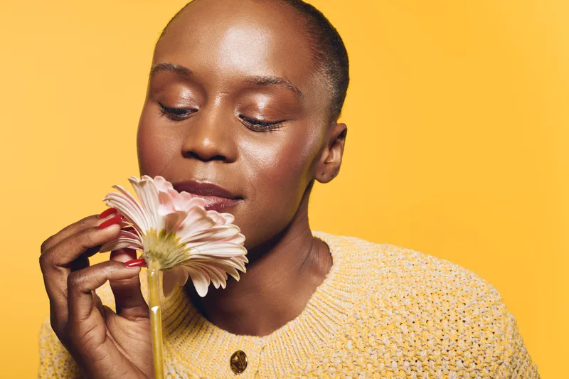 Close-up of a young African woman with dark skin holding a pink flower near her face. She wears a yellow sweater and poses against a yellow background. Concept of beauty and nature.