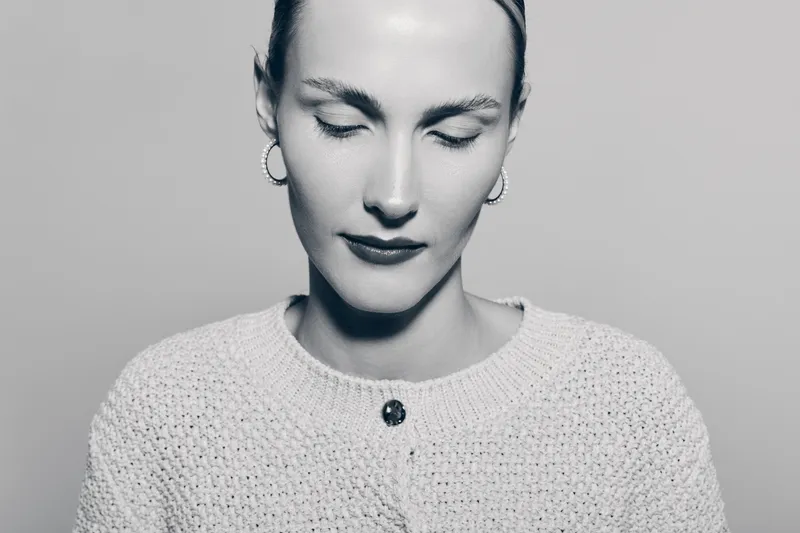 Close up of a calm Caucasian woman with pale skin and closed eyes wearing earrings and knitted sweater on neutral background.