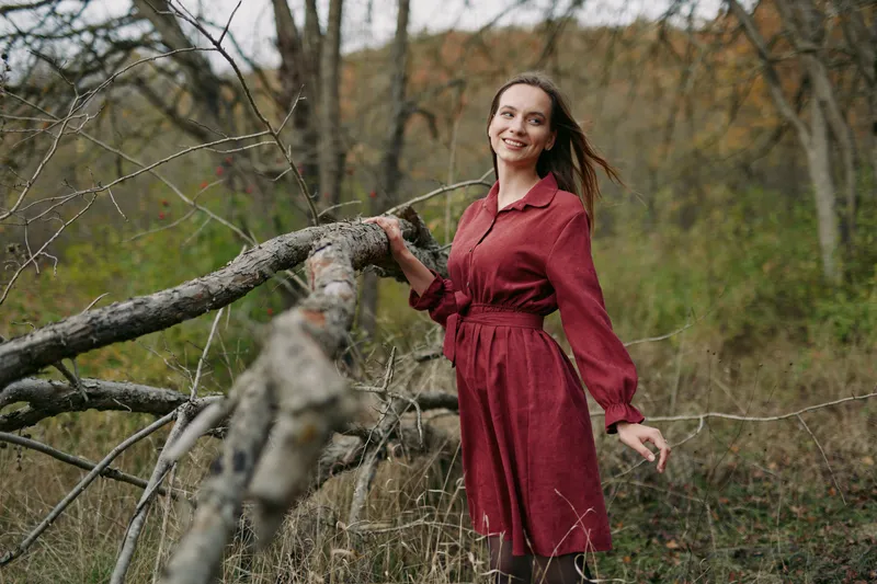 Authentic portrait of a woman in a burgundy dress leaning on a fallen branch in a quiet forest, conveying sincerity and natural light in a candid outdoor setting.