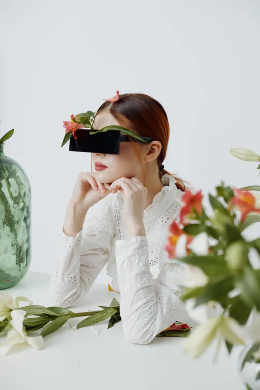 A young woman with long hair in a white blouse, wearing oversized sunglasses and floral accents, poses thoughtfully in a minimalist space with green and white tones, expressing calmness and creativit