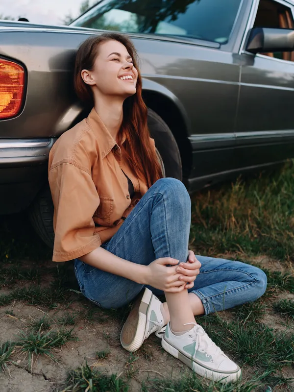 Young woman smiling joyfully while sitting beside a vintage car, wearing casual attire and enjoying a sunny day in nature.