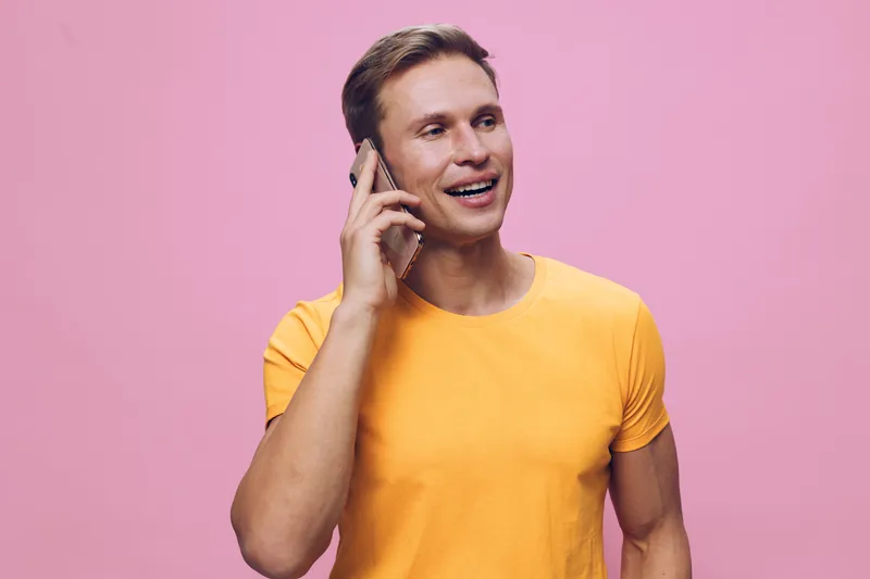 Smiling man using mobile phone talking on device with happy expression wearing casual yellow t-shirt isolated on pink background in studio. People lifestyle concept, communication, technology, positiv