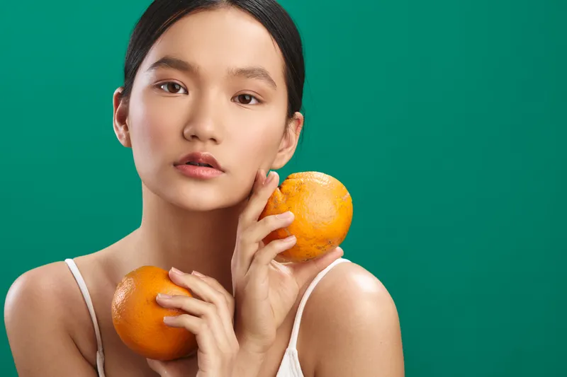 asian woman with a calm expression holds fresh oranges against a vivid green background, portrait style, clean studio lighting, minimal wardrobe, vibrant yet natural mood.
