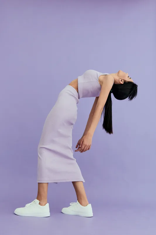 woman bending backward in lavender dress and white sneakers against light purple background, showcasing graceful motion and minimalist fashion style
