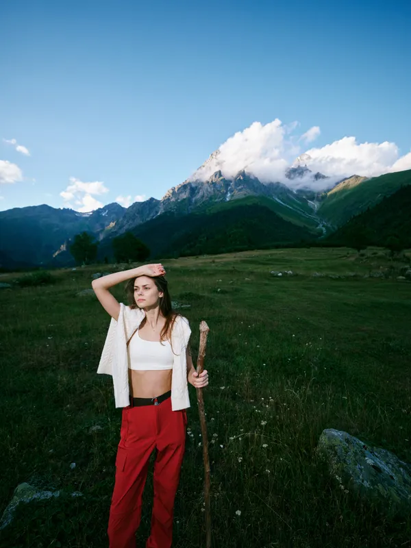 Hiking woman standing in a grassy meadow with a wooden staff, mountains in the distance under a blue sky, sunlit outdoors, casual attire, sense of adventure and exploration in nature