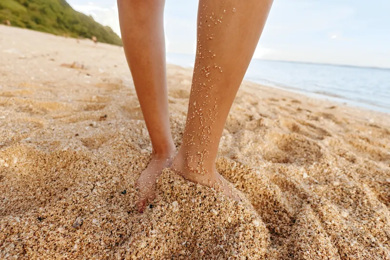 Close up of bare feet standing on sandy beach with grains of sand sticking to skin. Calm sea and blue sky in background create a peaceful summer atmosphere.