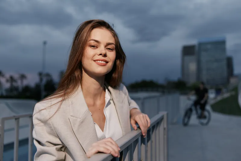 confident woman in beige blazer smiling outdoors in urban park with cloudy sky and modern buildings KEYWORD: confident woman smiling outdoors urban park beige blazer modern buildings evening light nat
