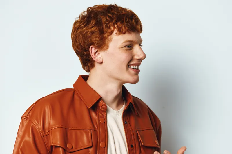 Young man with red hair wearing brown leather jacket and white shirt smiling and looking sideways against light background in casual style portrait.