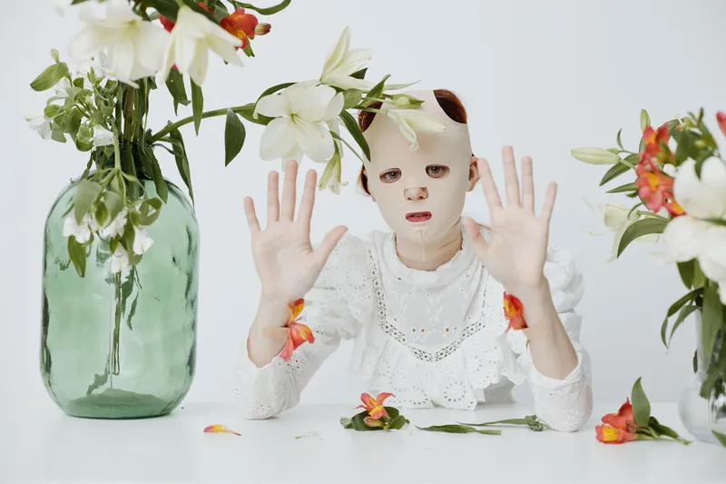 Unique conceptual portrait of a model with a mask in a floral setting, expressing emotions of curiosity and surrealism, featuring a mix of colors and textures