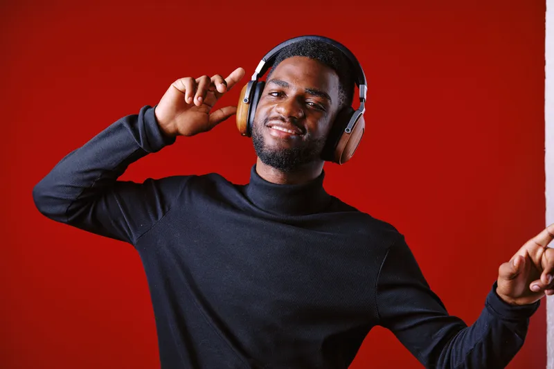 Young Black male enjoying music with headphones, expressing joy against a vibrant red background, stylish and modern outfit, perfect for music and lifestyle themes
