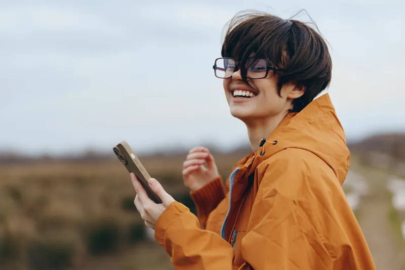 Woman lifestyle smartphone outdoors smiling glasses jacket portrait travel technology, orange jacket, outdoors smiling, holding smartphone, travel mood, soft daylight background, portrait
