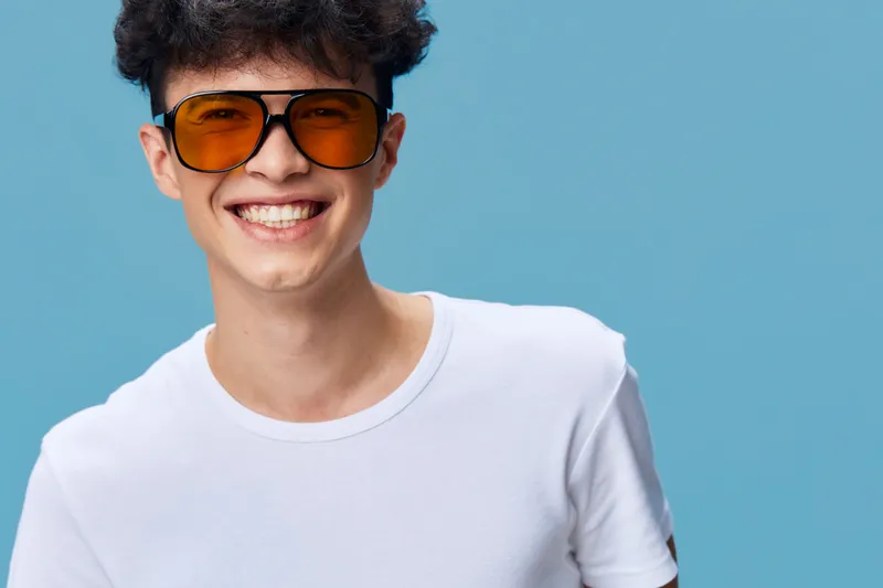 smiling man in white t-shirt wearing orange sunglasses with curly hair, joyful expression, isolated on blue background, casual style, youth culture