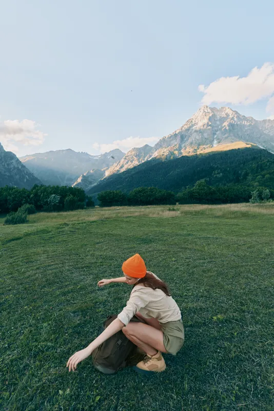 Woman outdoors, crouching in a field, mountain backdrop, orange beanie, casual top and shorts, natural light, relaxed pose, exploring nature, peaceful vibe