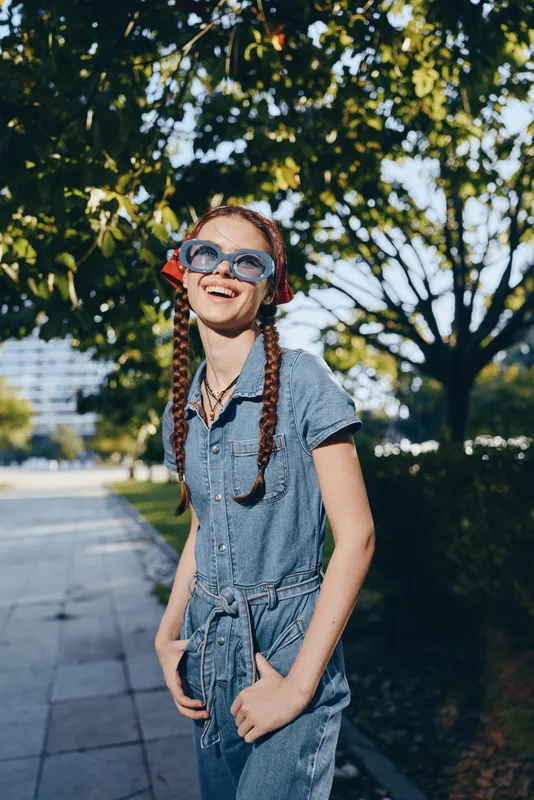 Lifestyle woman in modern boho-western denim street style smiling outdoors with braided hair and sunglasses in bright film color tones on a sunny day.