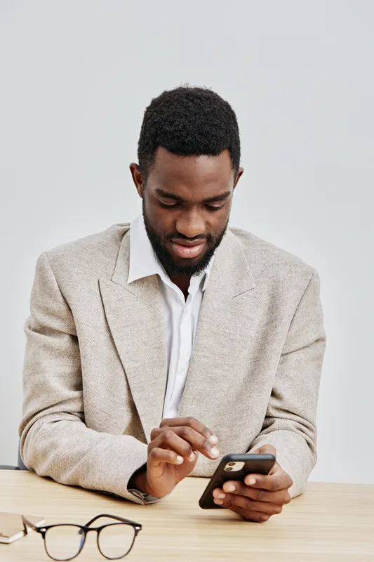Smiling man using smartphone, casual beige blazer and white shirt, modern workspace background, engaged and happy, technology and communication concept, lifestyle shot, vibrant colors, portrait orient