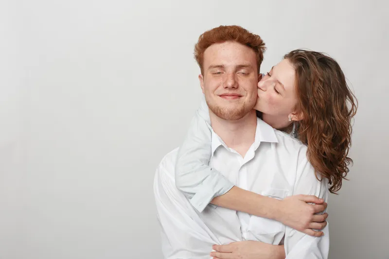 Happy young couple embraces and shares a tender moment, woman kissing man on cheek. Casual clothing and neutral background create warm, intimate, and loving relationship concept.