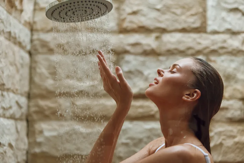 woman enjoying outdoor shower with water falling, relaxed expression, natural stone wall background, summer wellness concept