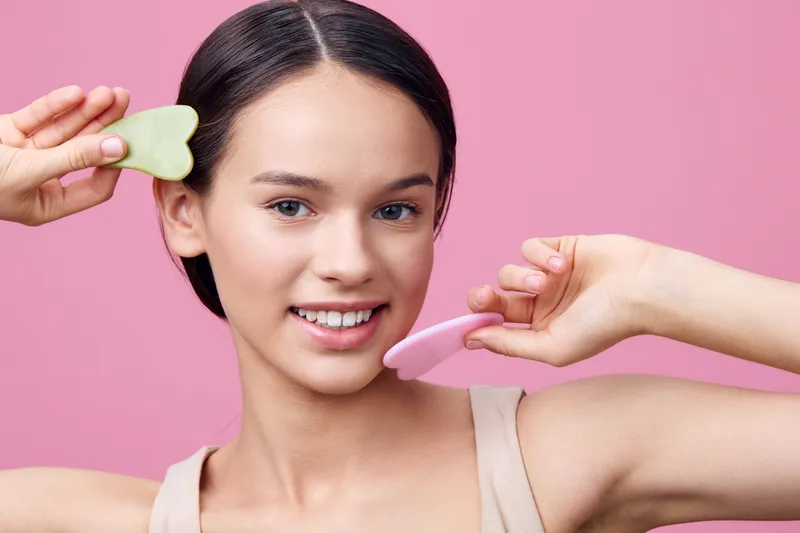 Young woman demonstrating facial massage techniques with jade and rose quartz tools on a pink background, showcasing healthy skincare and beauty routines