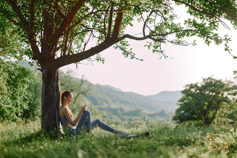 Young woman sitting under a tree in a lush green landscape, using a smartphone while enjoying nature s tranquility in the summer sun