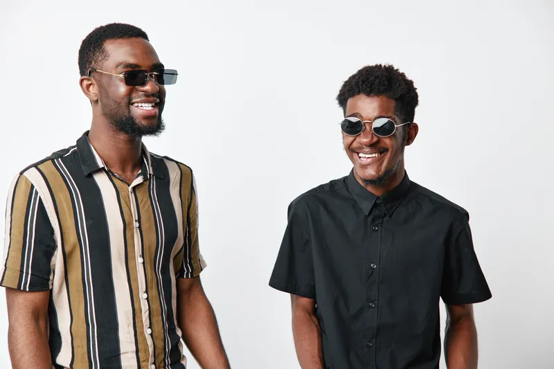 Two young black men with dark skin wearing sunglasses smiling and standing together in casual shirts on white background. Friendship, happiness and style with positive mood and fashion.
