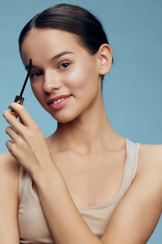 Young model applying mascara for a natural beauty look, showcasing skincare and makeup techniques against a soft blue background