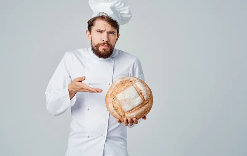 Шеф-повар в светлом костюме и головном уборе держит в руке круглый каравай хлеба