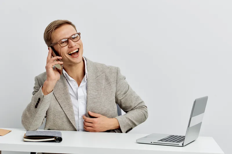happy young man wearing glasses talking on smartphone smiling in light gray blazer using laptop for remote work digital nomad concept modern workspace white background
