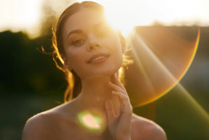 Young woman with glowing skin smiles serenely in natural sunlight, evoking warmth and beauty, surrounded by greenery and sunshine
