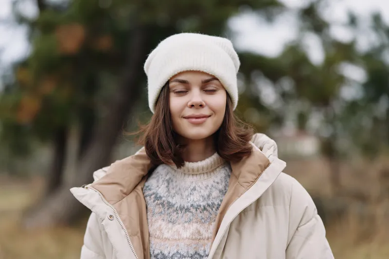 Woman in warm outerwear enjoys a calm outdoor moment, wearing a cozy knit sweater and a soft puffer coat with a beanie, basking in gentle autumn light and fresh park air.